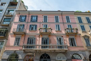 A multi-story building with a pink facade, multiple balconies with plants, and storefronts including 'Atelier VM' and 'Gioielli Valici'. - De Amicis' Nest - City Center (Milano)