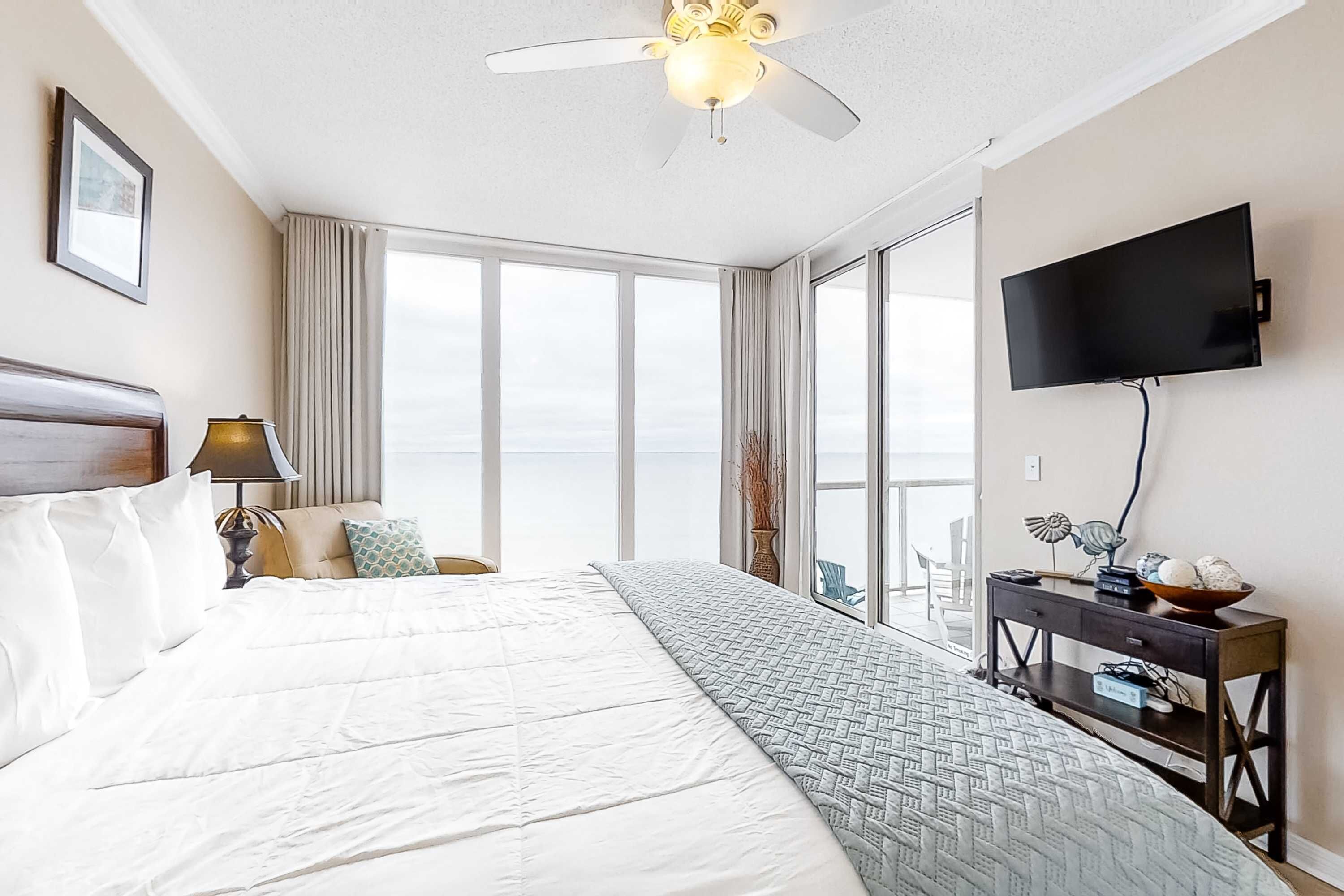 Quarto de hotel com uma cama grande, um ventilador de teto, uma televisão e vista para o céu através de amplas janelas.