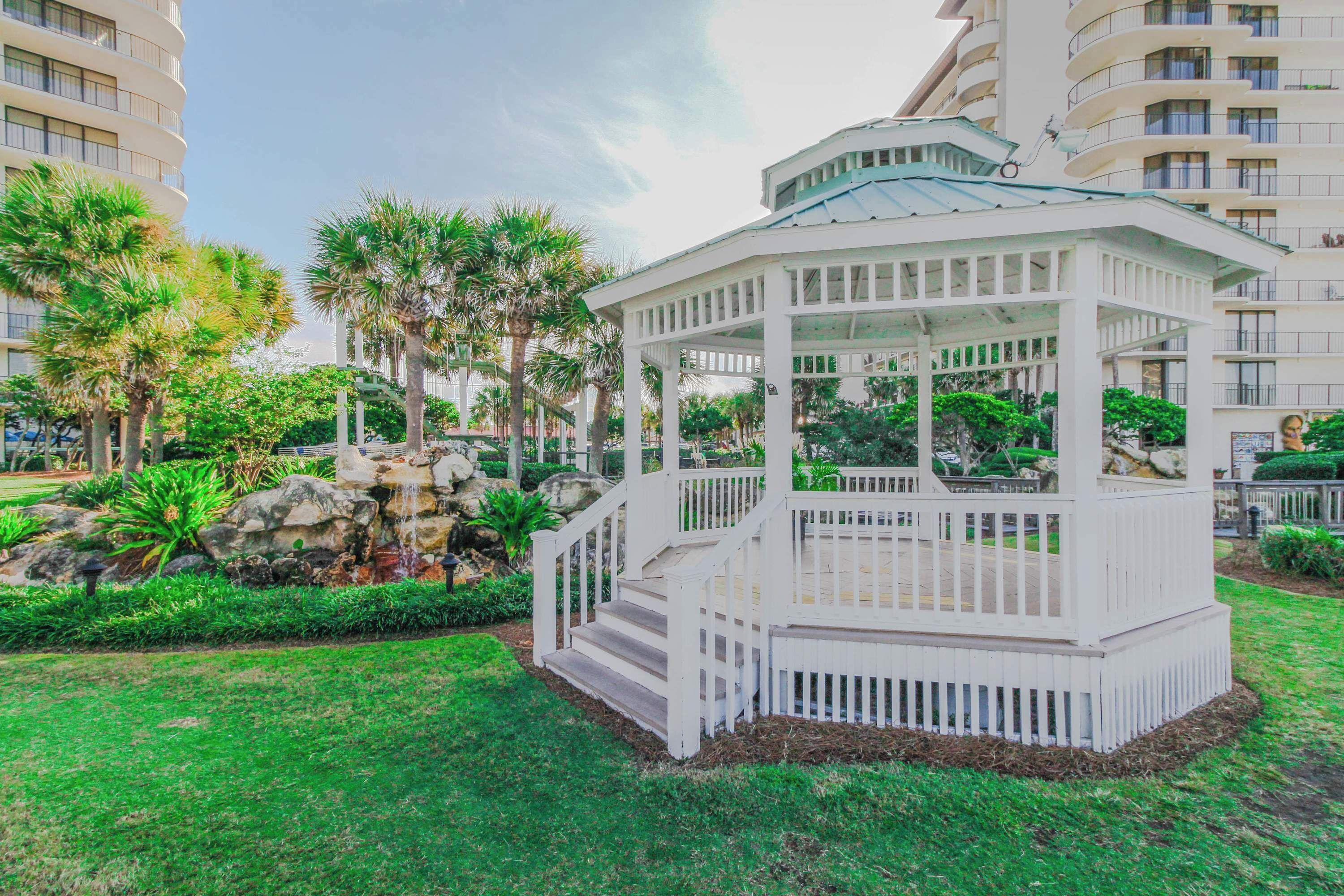 Un gazebo bianco con tetto e gradini, immerso nel verde tra l'erba e palme, in un'area curata con un edificio sullo sfondo.