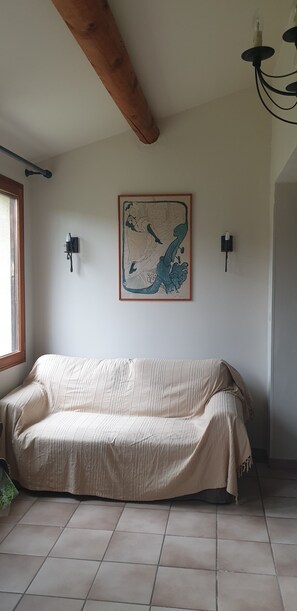 A room with a sofa, a wall-mounted painting, two wall sconces, a wooden ceiling beam, and a window. - Quiet 4-person vacation rental in the countryside, with amenities just a 5-minute walk away, a pool, and more (Maubec)