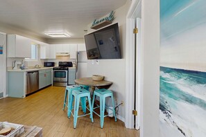 A kitchen with white cabinets, a microwave, an oven, and a sink. There are three turquoise stools around a round table. A painting of a seascape is on the wall. - Cozy coastal condo with shared pool and close to beach access (Panama City Beach)