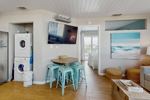 A living room with a washing machine, a TV, and a painting of the ocean. - Cozy coastal condo with shared pool and close to beach access (Panama City Beach)