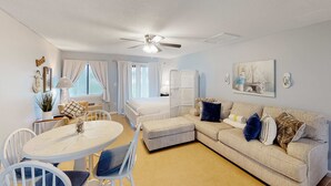 A well-lit, modern living room with a ceiling fan, a dining table with chairs, a sofa with cushions, and a large window with curtains.