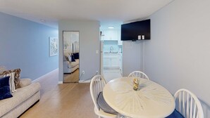 A living room with a sofa, a dining table with chairs, a TV mounted on the wall, and a kitchen visible through an open door.