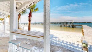 Un muelle en la playa con cubierta de madera y un área de estar techada con taburetes blancos.