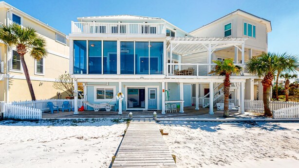 Una casa frente al mar de dos pisos, con una terraza de madera que conduce a la entrada, cerca viva blanca y palmeras.