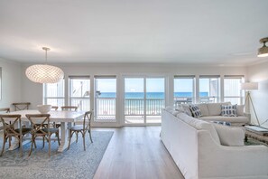 Een ruime woonkamer met een grote witte bank, een eettafel met stoelen en uitzicht op de oceaan door grote schuifdeuren.