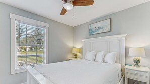 Une chambre avec un grand lit, un ventilateur de plafond, une fenêtre avec des stores, une table de chevet avec une lampe et un tableau au mur.