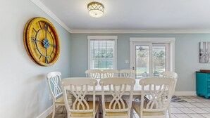 Un coin repas avec une table en bois et des chaises blanches, une grande horloge murale et une fenêtre avec des stores.