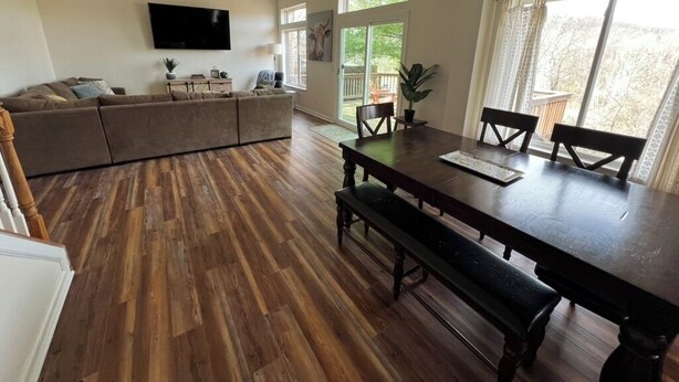 A spacious living area with a wooden floor, a dark wooden dining table with benches, a ceiling fan, and a flat-screen TV mounted on the wall.