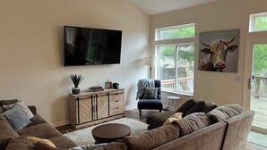 A living room with a sectional sofa, a flat-screen TV, a ceiling fan, a window with a view, and a painting of a cow.