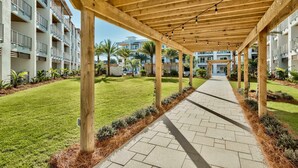 A covered walkway with wooden pillars and string lights, leading to a building with balconies and a well-maintained lawn. - Modern condo with pool, hot tub, gym, wrap-around balcony, & AC (Inlet Beach)
