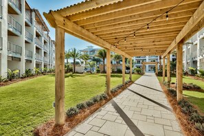 A covered walkway with wooden pillars and string lights, leading to a building with balconies and a well-maintained lawn. - Modern condo with pool, hot tub, gym, wrap-around balcony, & AC (Inlet Beach)