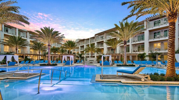 A pool area with multiple lounge chairs and palm trees, surrounded by multi-story buildings with balconies. - Modern condo with pool, hot tub, gym, wrap-around balcony, & AC (Inlet Beach)