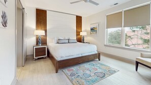 A bedroom with a bed, bedside tables, a ceiling fan, a window with blinds, and a view of trees outside. - Modern condo with pool, hot tub, gym, wrap-around balcony, & AC (Inlet Beach)