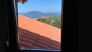 Vista a través de una ventana de un edificio con tejado de tejas rojas, cielo azul despejado y una costa lejana con barcos y una pequeña isla.