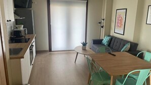 Un espacio habitable compacto con kitchenette, una zona de comedor con mesa y sillas, un sofá y una puerta corrediza que da a un balcón.