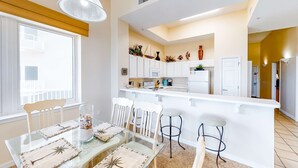 A modern kitchen with white cabinets, a central island, and a dining area with a glass table and white chairs. - First-Floor Gulf Front Condo with Shared Pool & Hot Tub (Navarre Beach)