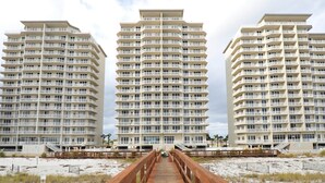 A beachfront hotel with multiple floors, a wooden boardwalk leading to the entrance, and a sandy beach in the foreground. - First-Floor Gulf Front Condo with Shared Pool & Hot Tub (Navarre Beach)
