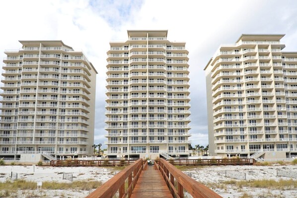 A beachfront hotel with multiple floors, a wooden boardwalk leading to the entrance, and a sandy beach in the foreground. - First-Floor Gulf Front Condo with Shared Pool & Hot Tub (Navarre Beach)