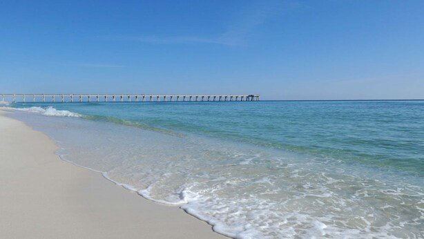 A beach with clear blue water and a pier extending into the distance. - First-Floor Gulf Front Condo with Shared Pool & Hot Tub (Navarre Beach)