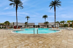 Piscina com palmeiras e espreguiçadeiras, cercada por edifícios com chão ladrilhado.