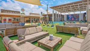 Un espace extérieur aménagé avec des canapés et des tables, un grand écran diffusant des événements sportifs, et des points de vente de nourriture à proximité.