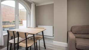 A modern living room with a dining table and chairs, a sofa, and a large window with a view of a brick wall and greenery outside. - Charming 2-bedroom Apartment in Central Kingston Upon Thames (Kingston Upon Thames)