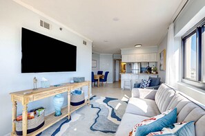 Een moderne woonkamer met een flatscreen televisie, een houten console en een grijze bank met blauwe kussens, en een keuken op de achtergrond.