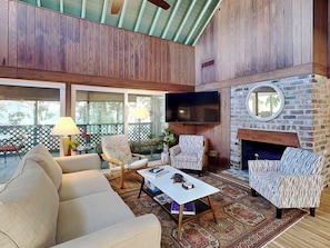 A spacious living room with a high ceiling, wooden paneling, a large TV, a stone fireplace, and a view through large windows. - 6 7th Street | Private Pool, Walk to Beach (Tybee Island)