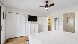 Un dormitorio con una cama grande, un armario blanco, un ventilador de techo y un televisor en la pared.