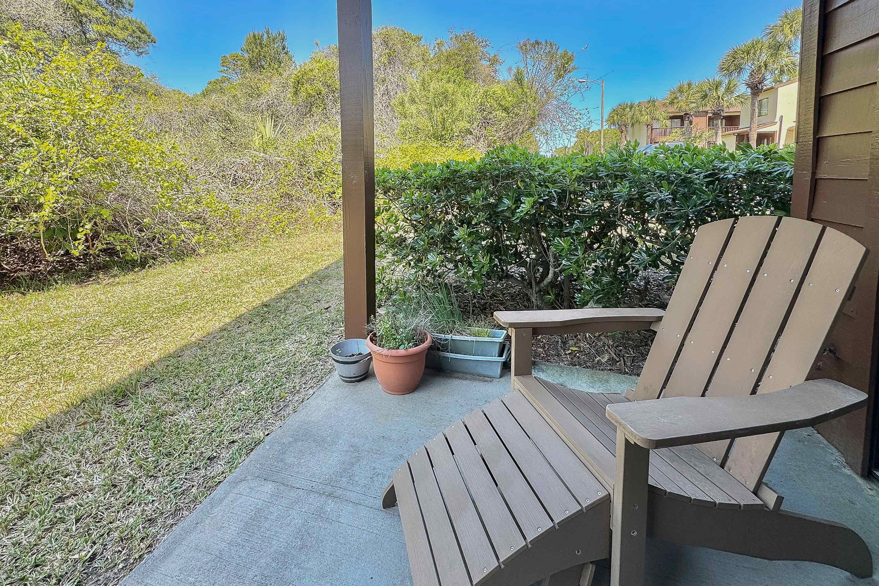 A wooden deck with a chair and a small table, surrounded by a garden and a hedge.