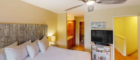 A bedroom with a bed, a ceiling fan, a TV on a dresser, and a bathroom visible through an open door.