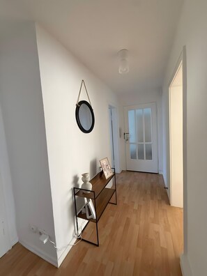 A hallway with a white wall, a wooden floor, a door with frosted glass panels, a small table with a vase and a picture frame, and a round mirror hanging on the wall. - Beautiful Apartment Opposite the Train Station (Kassel)