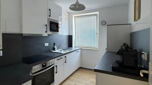 A modern kitchen with white cabinets, a black countertop, a microwave, a stove, and a window with blinds. - Beautiful Apartment Opposite the Train Station (Kassel)