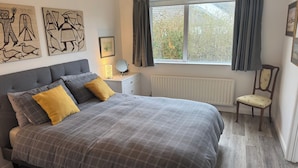 Een slaapkamer met een bed, nachtkastje, spiegel, stoel, raam met gordijnen en een kunstwerk aan de muur.