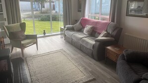Een woonkamer met een bank, een fauteuil, een salontafel en een groot raam met gordijnen.