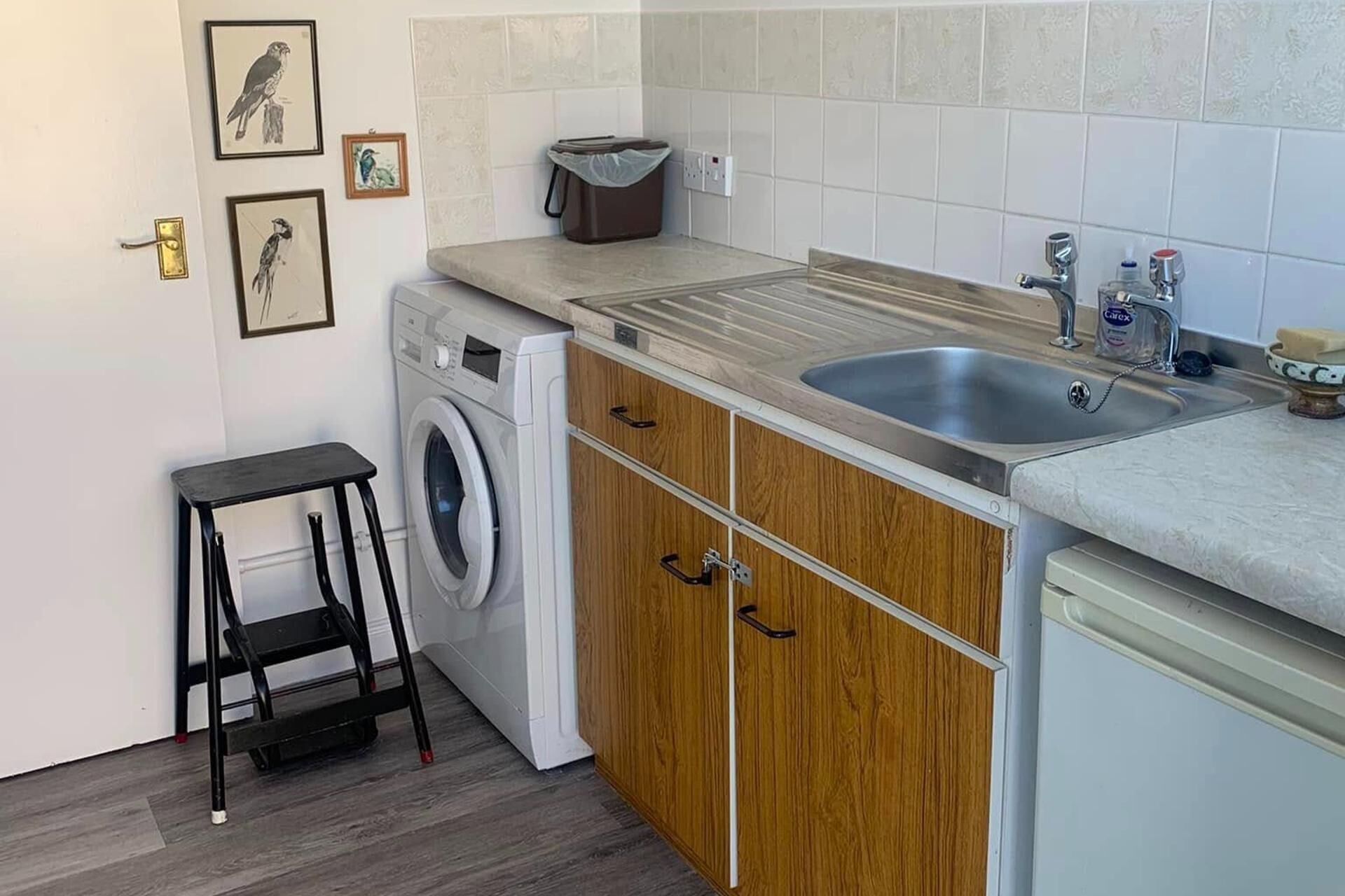 A compact kitchen with a washing machine, wooden cabinets, a sink, and a small stool.