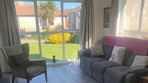 Een woonkamer met een groot raam, een groene fauteuil, een grijze bank met gekleurde kussens en een klein houten tafeltje.