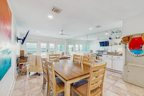 A spacious dining area with a wooden table and chairs, a kitchen with white cabinets, and a large window providing natural light. - Beautiful beachfront townhome, large deck and beach access (Panama City Beach)