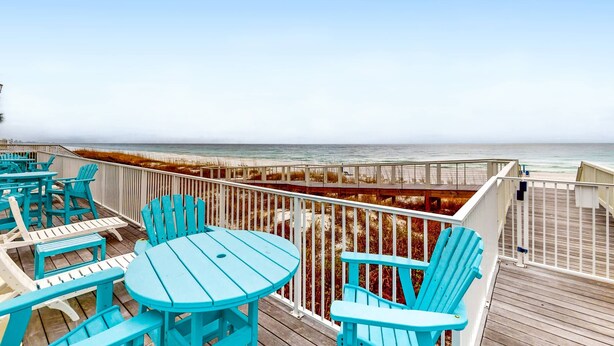 Een houten terras met turquoise stoelen en tafels, met uitzicht op een strand.
