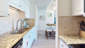 Eine schmale Küche mit Granit-Arbeitsplatten, weißen Schränken und einer Ziegelwand als Rückwand. Es gibt einen Essbereich mit Tisch und Stühlen sowie einen Messerblock auf der Arbeitsplatte.