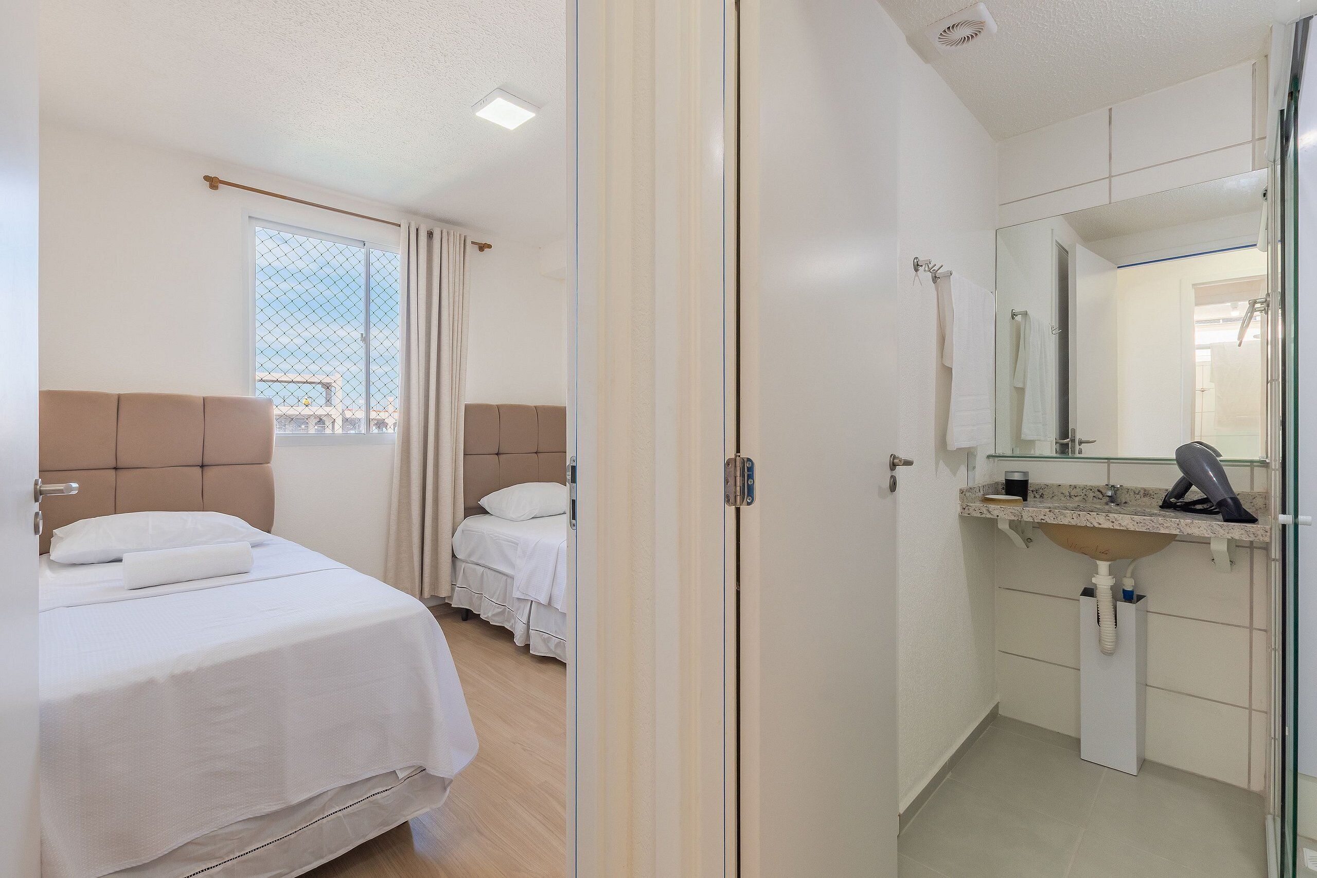 Una habitación de hotel con cama, mesita de noche, ventana con cortinas, baño con lavamanos y espejo, y zona de ducha.