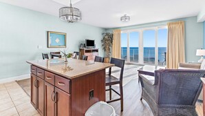 A modern kitchen with a central island, dining area with chairs, a TV, and a view of the ocean. - Beachfront condo with access to 5 pools, hot tub, fast WiFi, patio, washer/dryer (Panama City Beach)