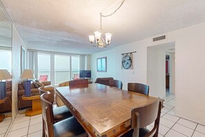 A spacious dining area with a large wooden table, several chairs, a chandelier, and a television in the background. - Bright waterfront condo in a gated resort with pools, tennis courts, & gym (Panama City Beach)