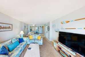 A living room with a sofa, coffee table, and a TV. There is a kitchen in the background. - Beachfront condo with unspoiled Gulf views, pool, & direct beach access (Panama City Beach)