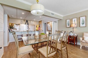 A dining area with a glass table and chairs, a chandelier, and framed artwork on the walls. - Gorgeous beachfront condo, shared & and beach access (Panama City Beach)