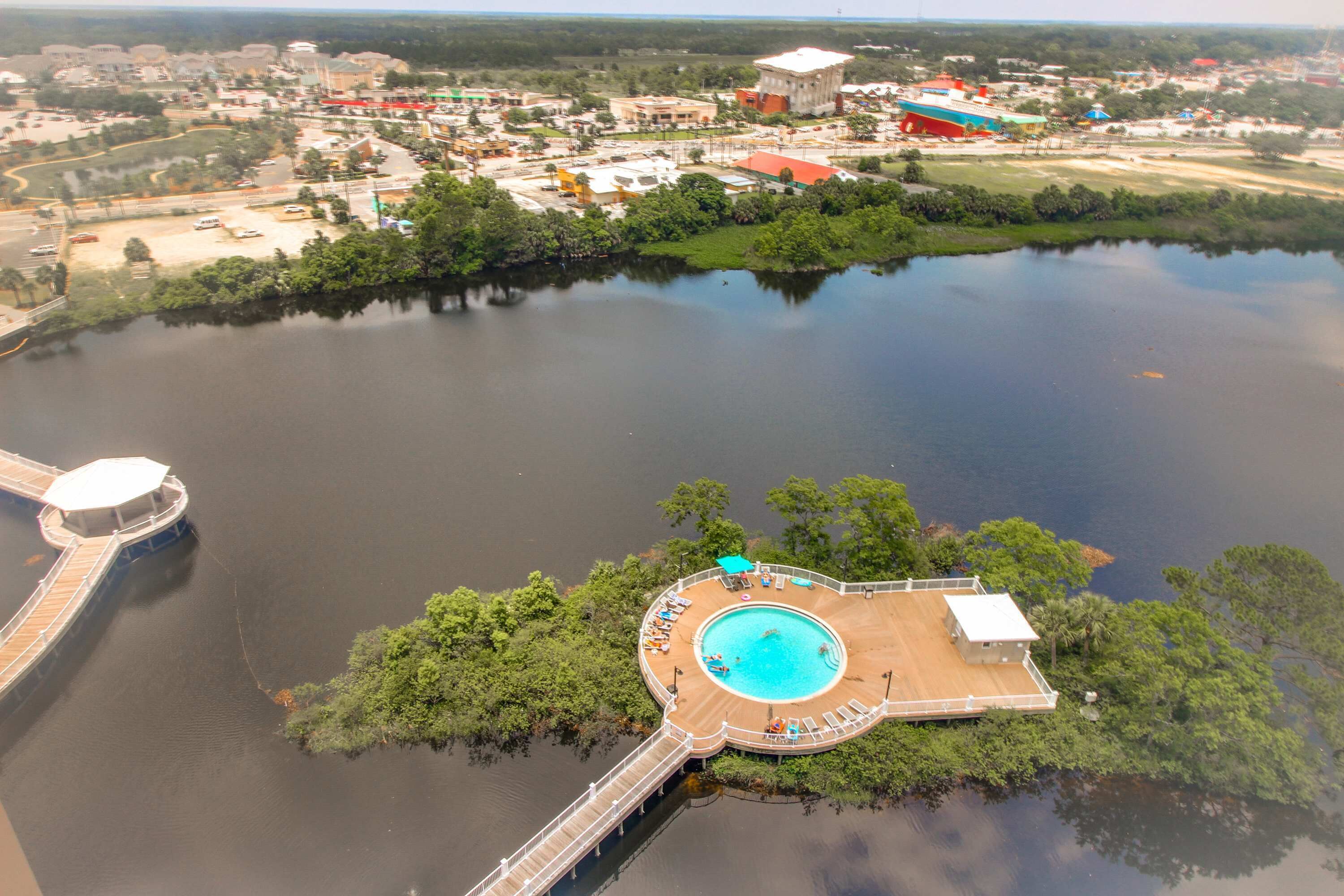 An aerial view of a lake with a circular swimming area surrounded by a wooden deck, a bridge, and a small building.