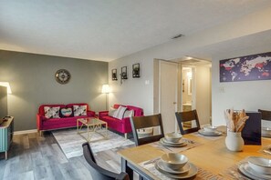 Een eetgedeelte met een houten tafel, gedekt voor vier personen, omringd door stoelen. Een rode bank met decoratieve kussens staat tegen de muur, en een klein bijzettafeltje met een lamp is aan de linkerkant te zien.