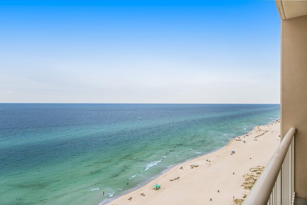 Blick auf einen Strand vom Balkon aus, mit klarem, blauen Wasser und einem sandigen Ufer, sowie einigen Sonnenschirmen.